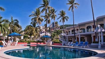 Zona de piscina con sillones de descanso y sombrillas, rodeada de palmeras y un edificio con ventanas arqueadas.