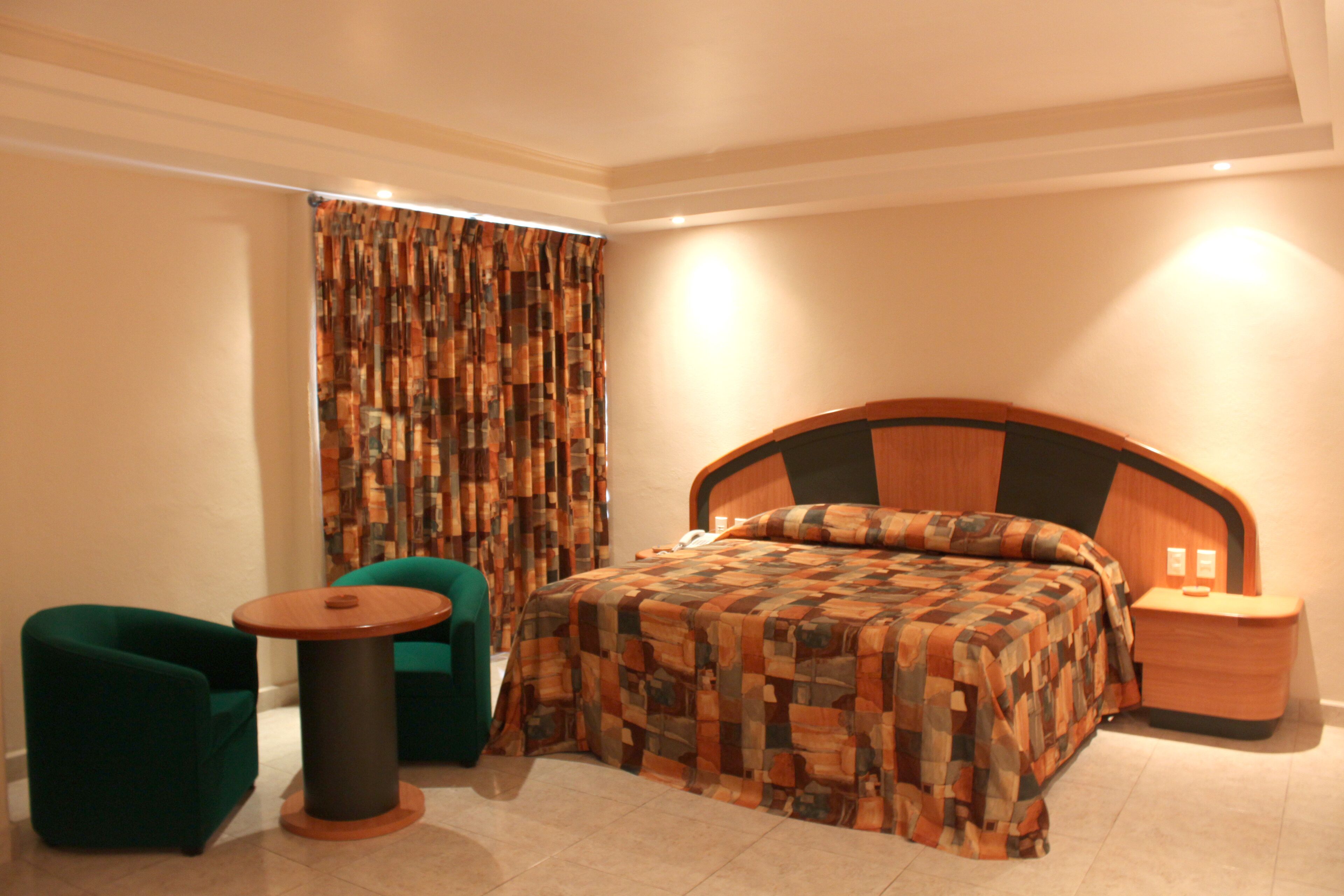 Un dormitorio con una cama grande, una mesita redonda pequeña, dos sillones verdes y una cortina con diseño geométrico.