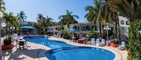 A pool area with lounge chairs and umbrellas, surrounded by palm trees and a building with arched windows.