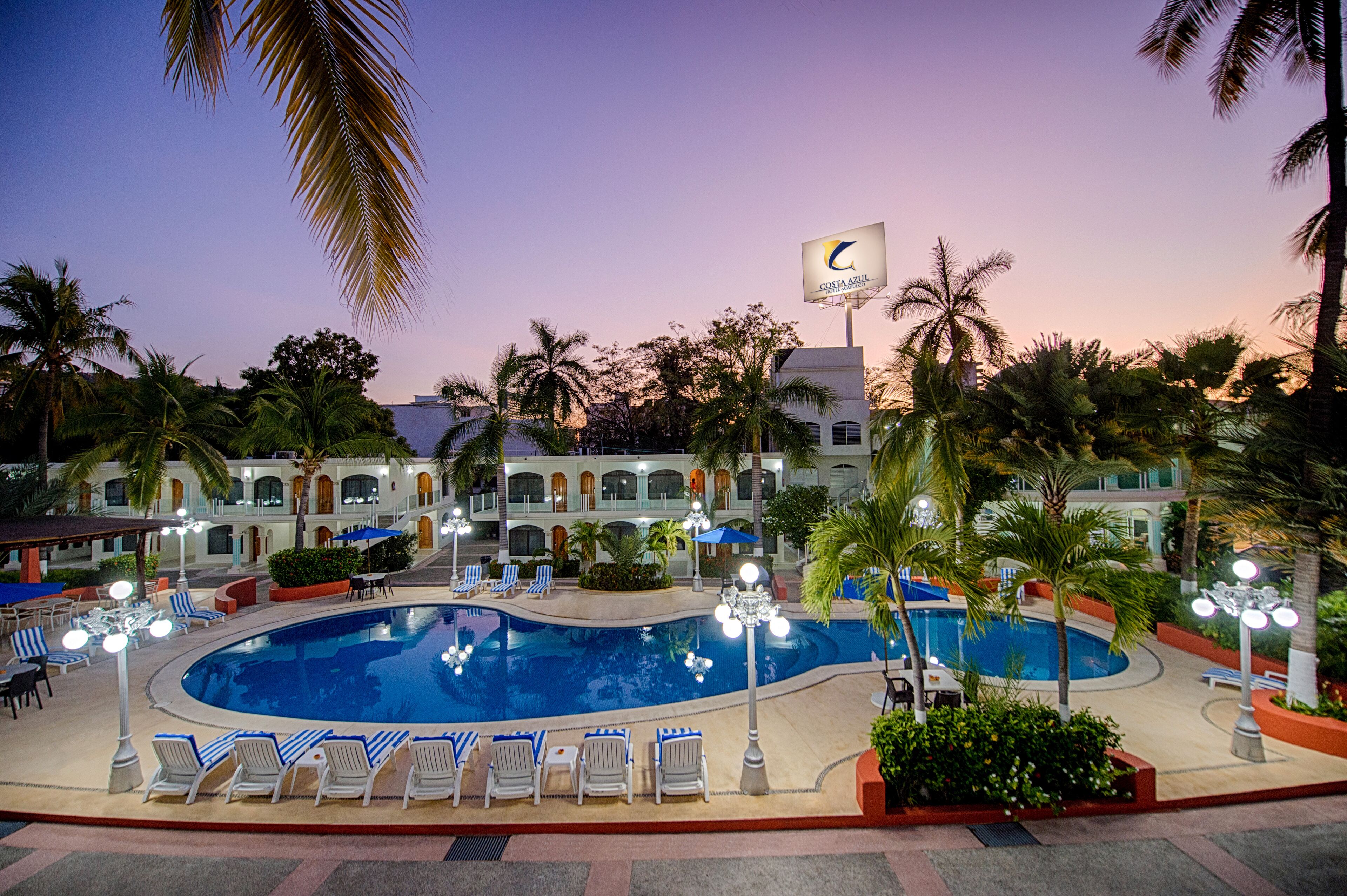 A resort pool area with lounge chairs, palm trees, and a building with a flag.