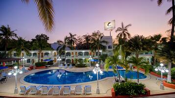 A resort pool area with lounge chairs, palm trees, and a building with a flag.