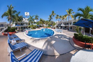 Un área de piscina en un complejo turístico con sillones reclinables, palmeras y un cielo azul despejado.