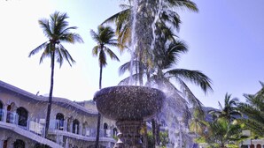 A fountain with water jets in a circular basin, surrounded by palm trees and a building with arched windows.