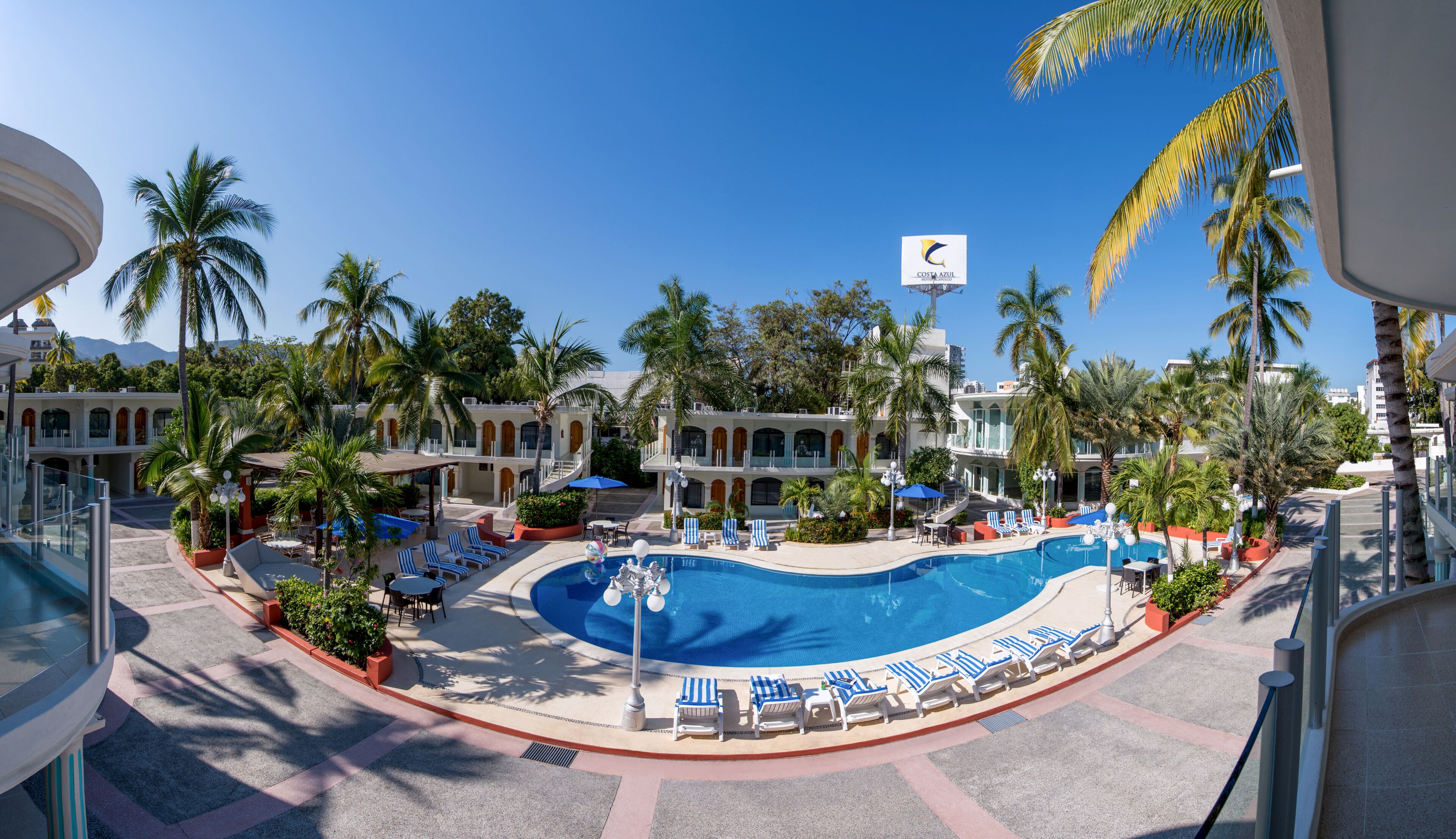 Ett semesterparadis med en central pool, solstolar, palmer och en klarblå himmel.
