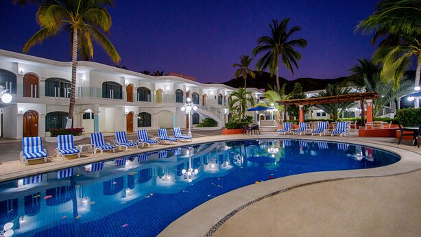 A swimming pool with blue and white striped lounge chairs, palm trees, and a building with arched windows and a covered patio area.