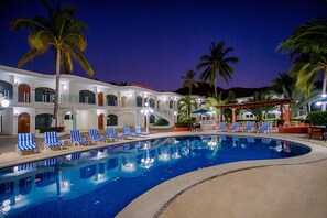 A swimming pool with blue and white striped lounge chairs, palm trees, and a building with arched windows and a covered patio area. - Hotel Costa Azul (Acapulco)