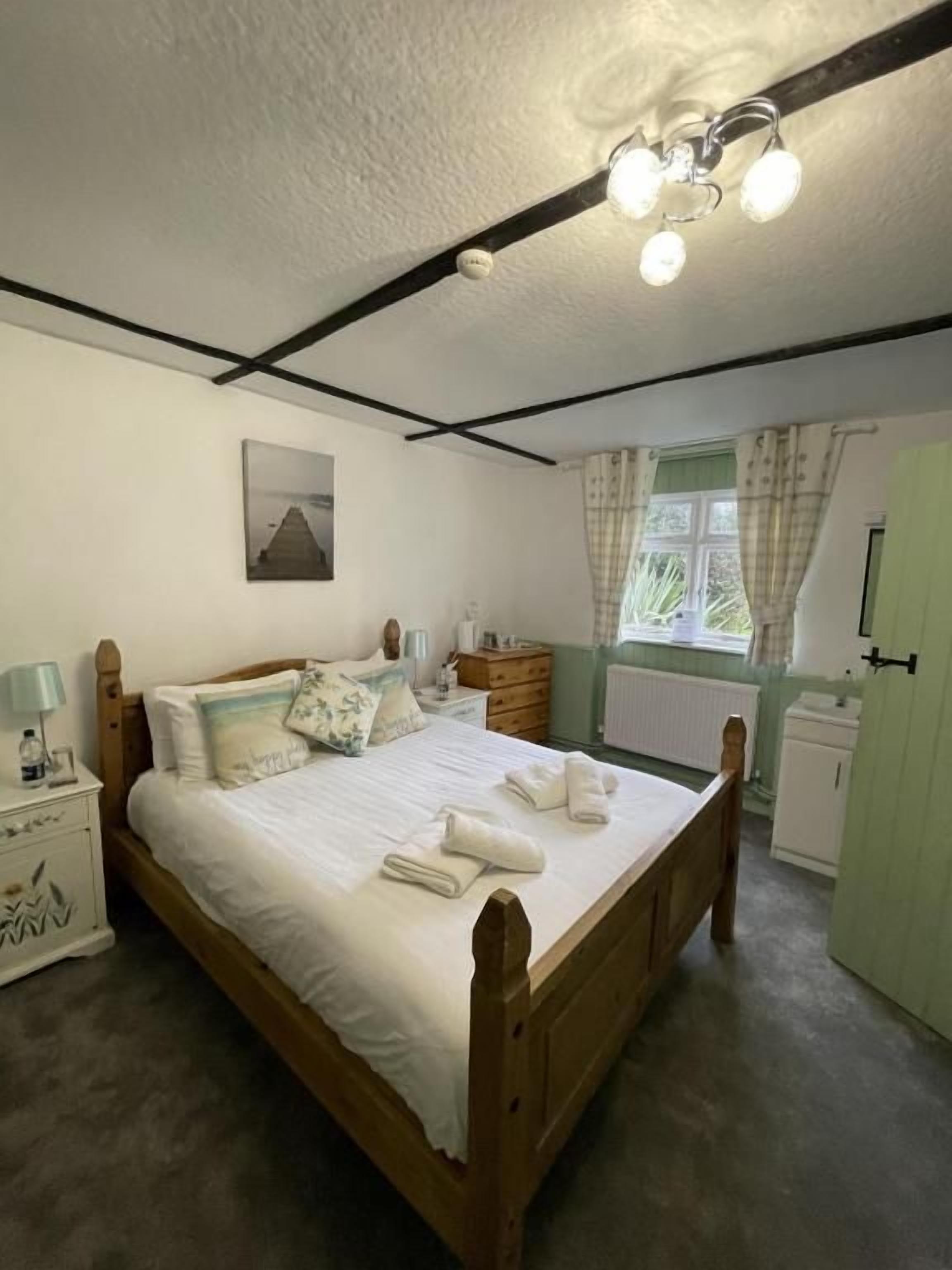 Ein Schlafzimmer mit einem hölzernen Bett, Nachttischen, einem Beistelltisch, einem Fenster mit Vorhängen und einer Heizung.