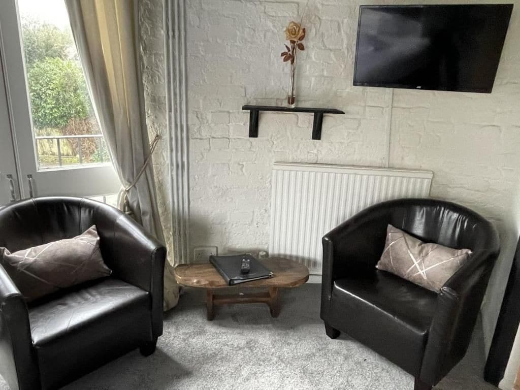 Une pièce comprenant deux fauteuils en cuir, une petite table en bois, un radiateur, un téléviseur fixé au mur et une fenêtre avec des rideaux.