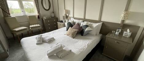 A neatly made bed with towels, a bedside table with a lamp, a chair, and a window with curtains.