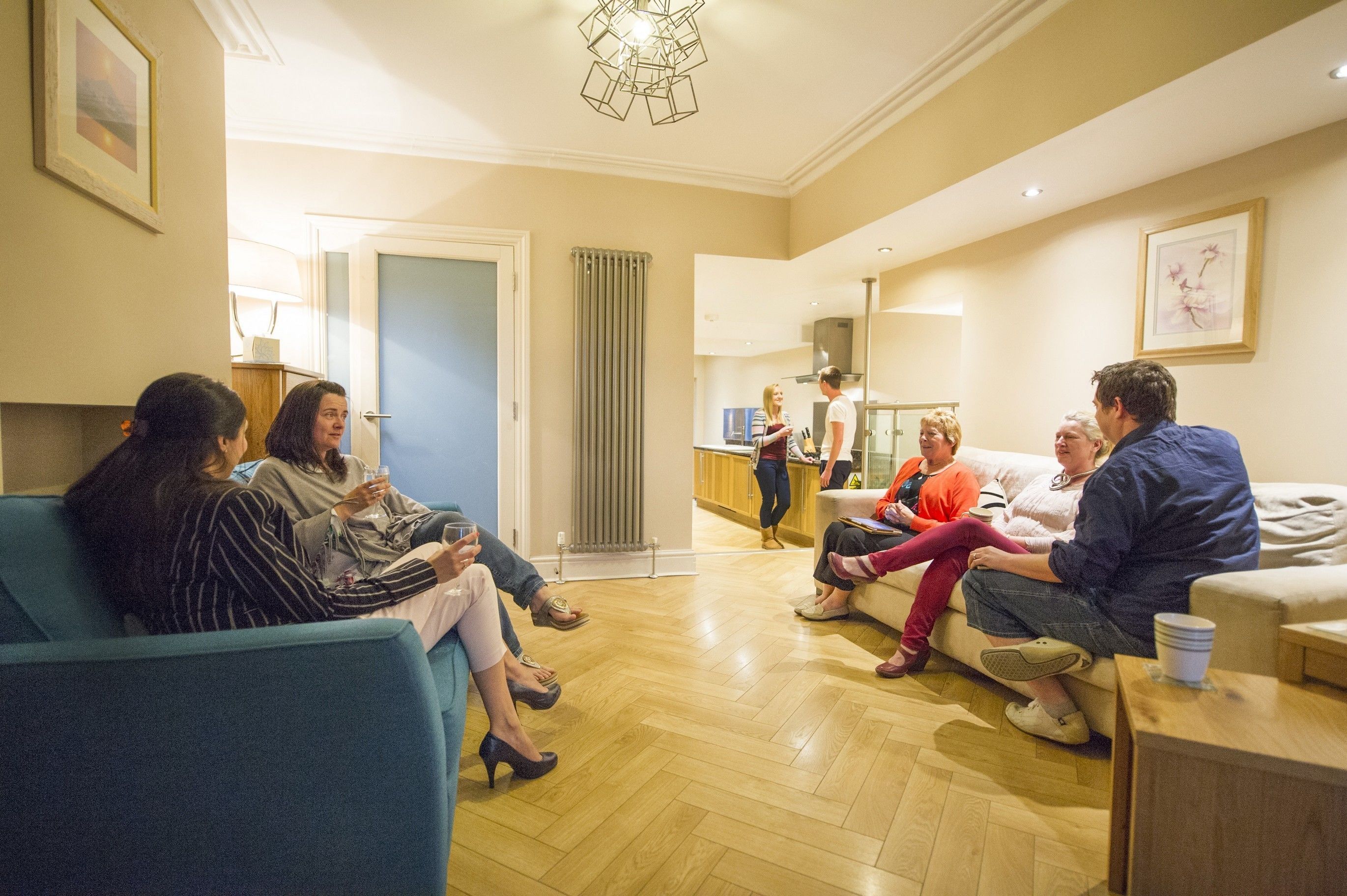 Un grupo de personas sentadas en un sofá azul en una habitación bien iluminada con suelo de madera, con una zona de cocina al fondo y un cuadro en la pared.