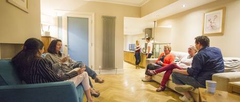 Un grupo de personas sentadas en un sofá azul en una habitación bien iluminada con suelo de madera, con una zona de cocina al fondo y un cuadro en la pared.