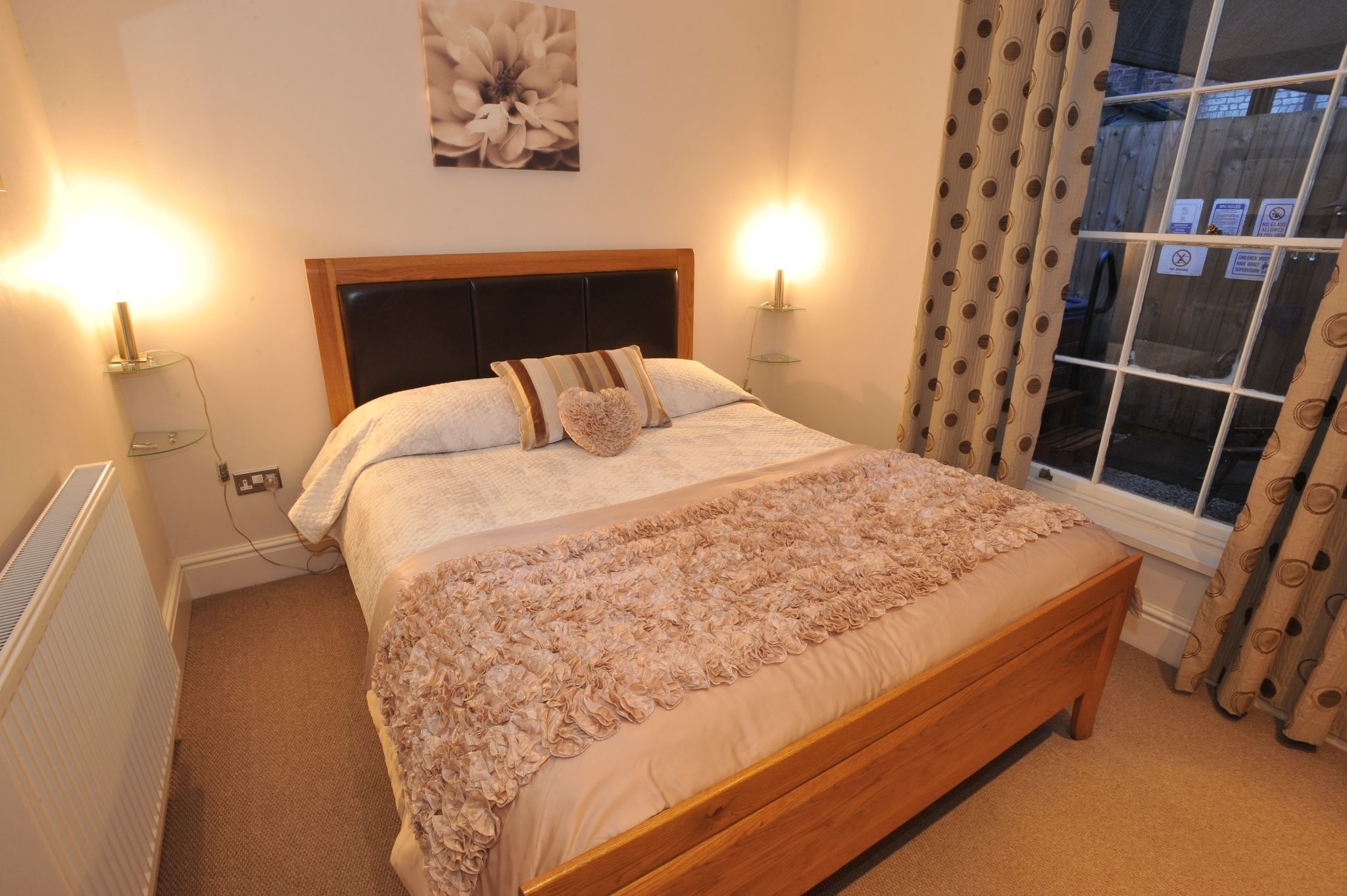 A neatly made bed with a wooden headboard, two bedside lamps, a floral wall art piece, and a window with curtains.
