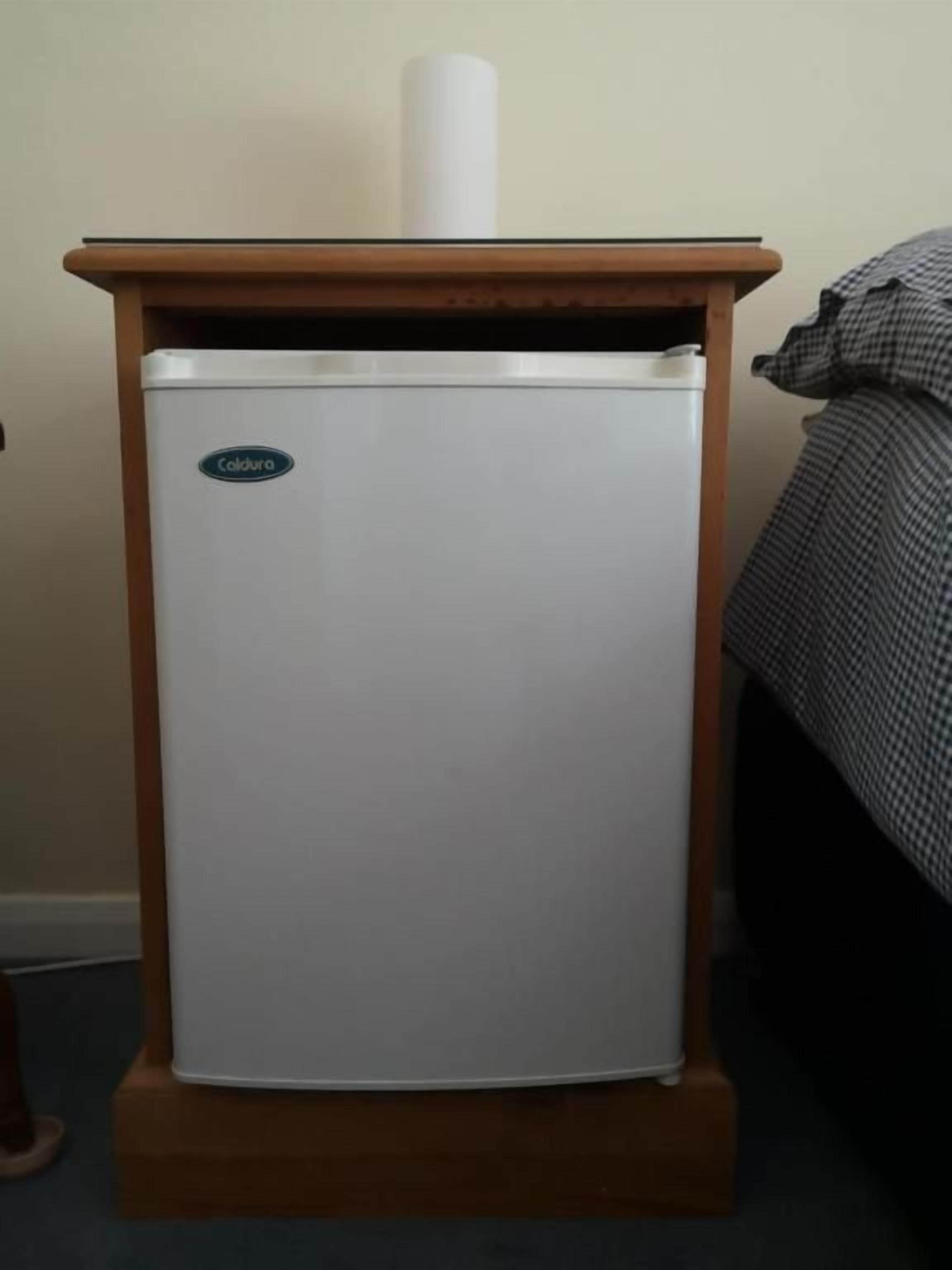 Un refrigerador compacto con base de madera, situado en una habitación con cama y pared.