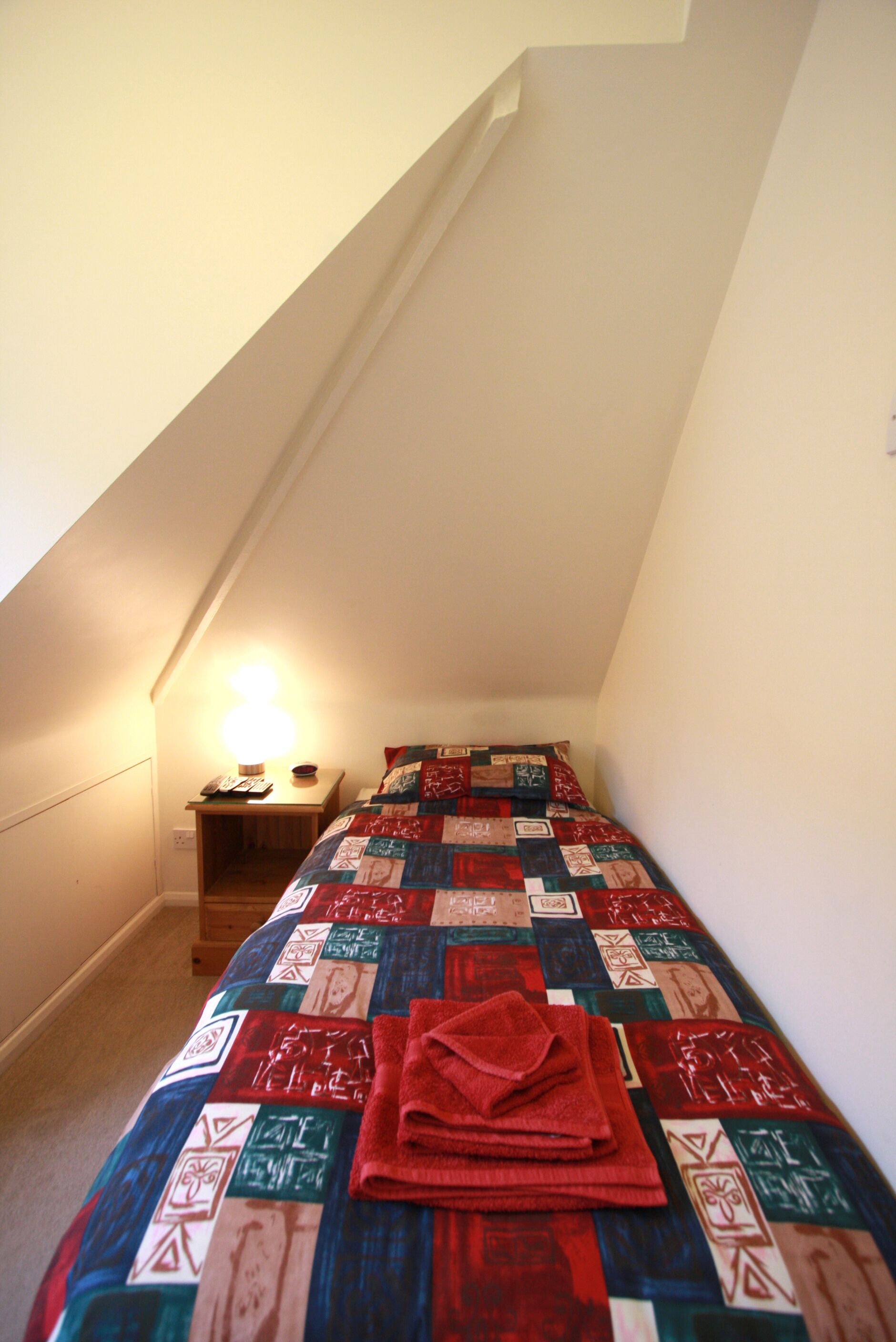 Une petite pièce étroite avec un plafond en pente, un lit recouvert d’une couverture colorée, une table de chevet avec une lampe et un luminaire fixé au mur.