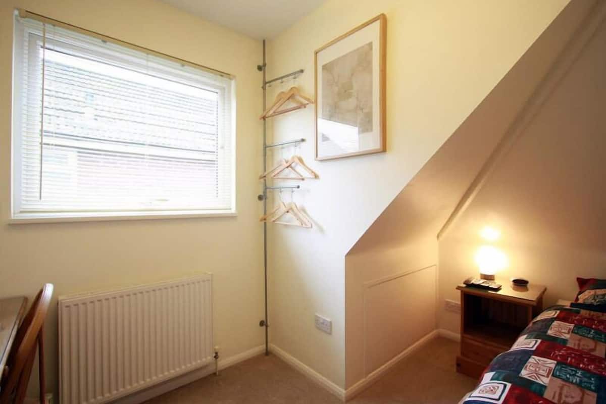A bedroom with a window, radiator, wooden hanger rack, framed picture, and a bedside table with a lamp.