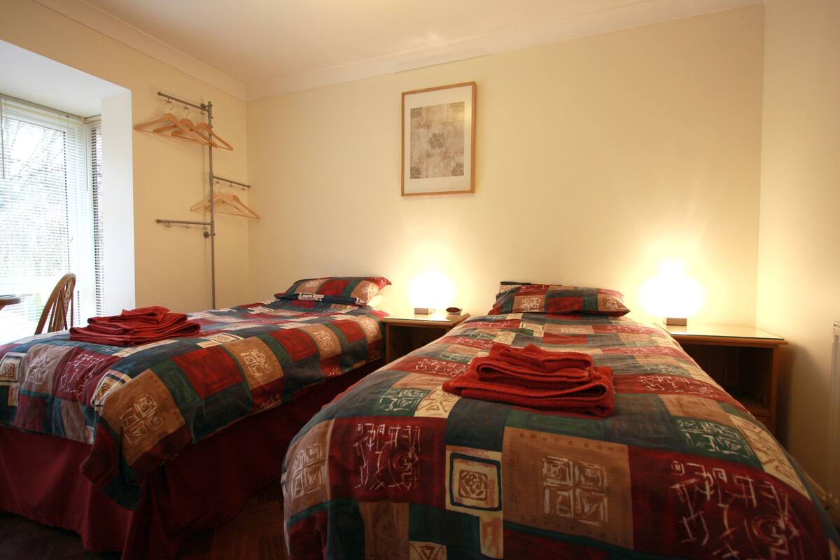 A hotel room with two beds, each with a quilted bedspread and a red towel on top. There is a framed picture on the wall above the beds and a window with blinds to the left.