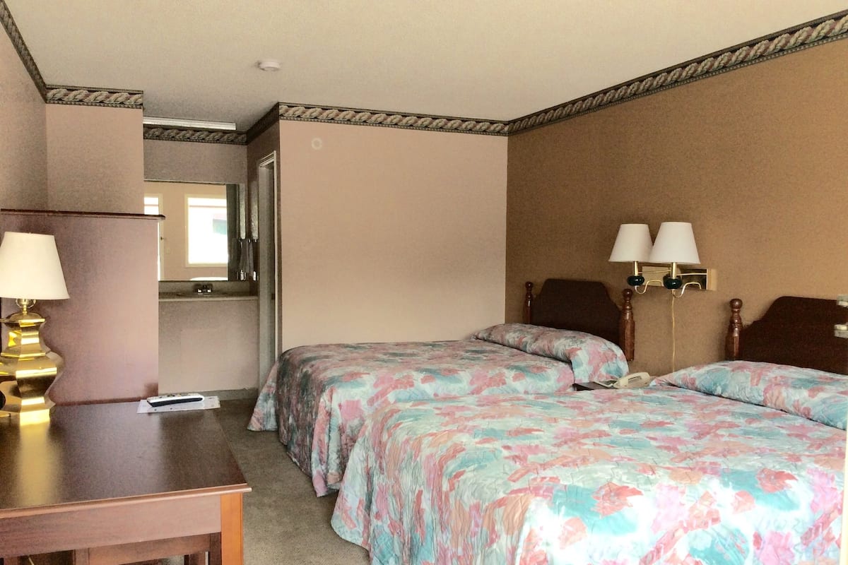 Quarto de hotel com duas camas, uma escrivaninha, um abajur e um espelho.