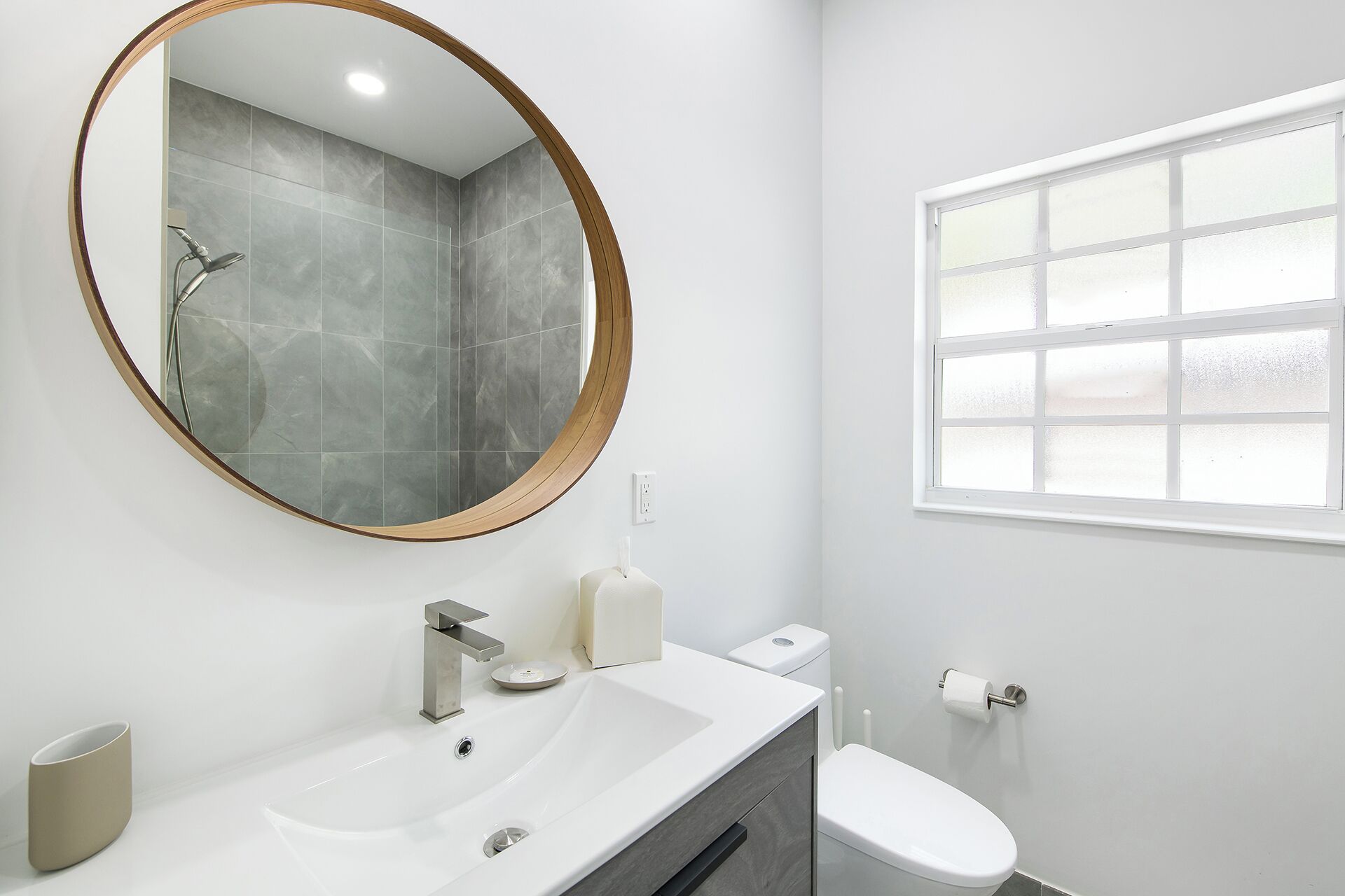 A bathroom with a round mirror, a sink, a toilet, and a window.