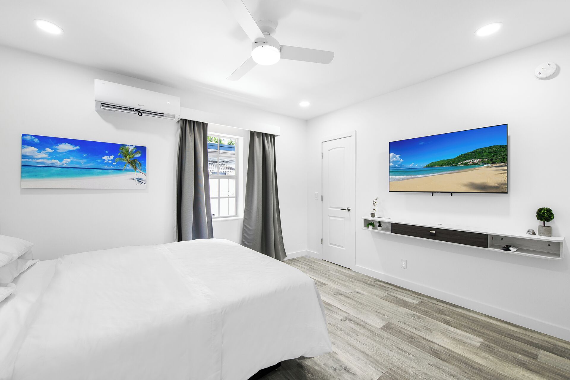 A bedroom with a bed, a ceiling fan, an air conditioning unit, a window with curtains, and two wall-mounted TVs displaying beach scenes.