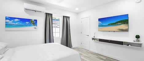 A bedroom with a bed, a ceiling fan, an air conditioning unit, a window with curtains, and two wall-mounted TVs displaying beach scenes.