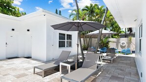 Eine Terrassenfläche mit Liegestühlen, einem Tisch und Sonnenschirmen. Zwei Türen und ein Fenster sind sichtbar.