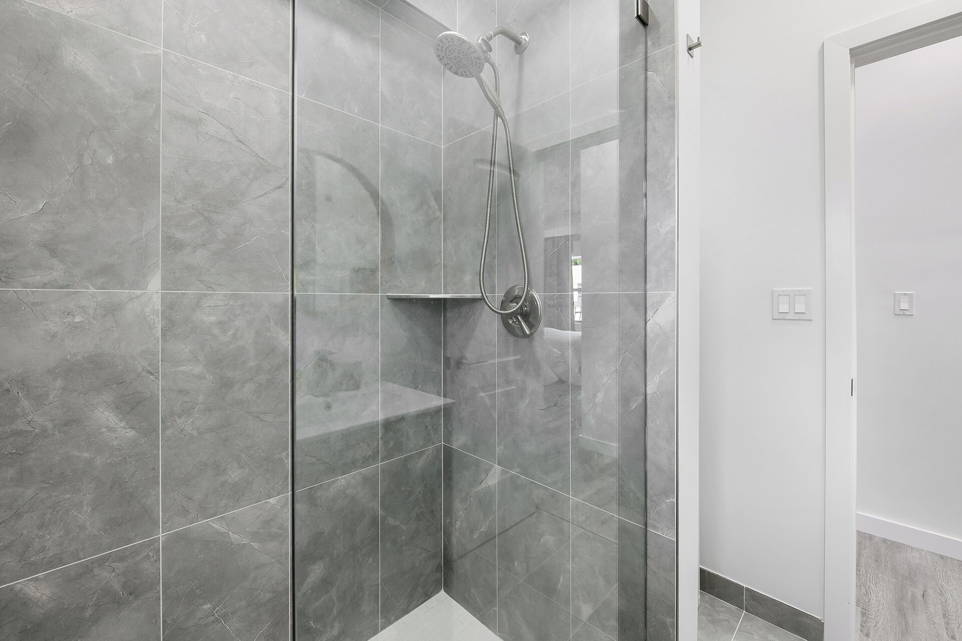 A modern bathroom with a glass-enclosed shower, grey marble tiles, and a white door.