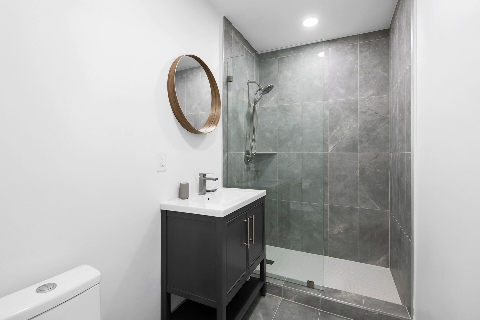 Een moderne badkamer met een witte wastafel, donker kastje en een douche met grijze tegels.