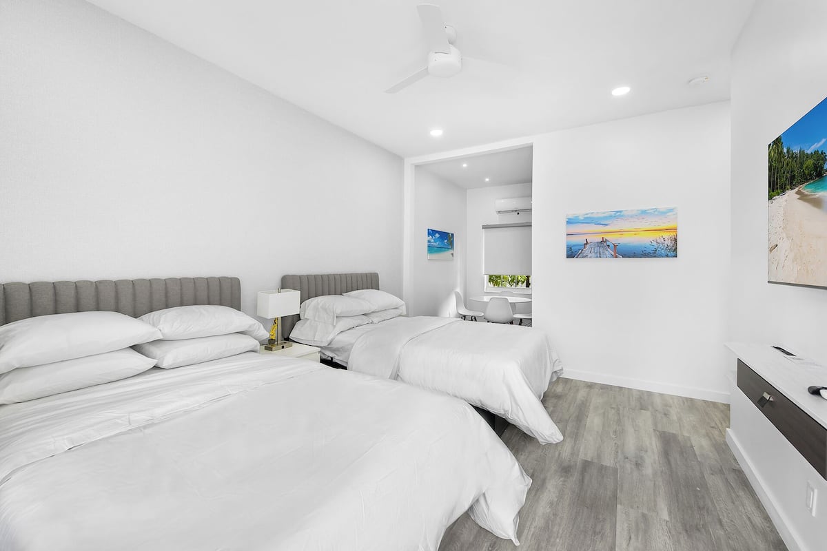 Habitación de hotel moderna con dos camas, ventilador de techo, un cuadro de una playa y vista a la playa desde la ventana.