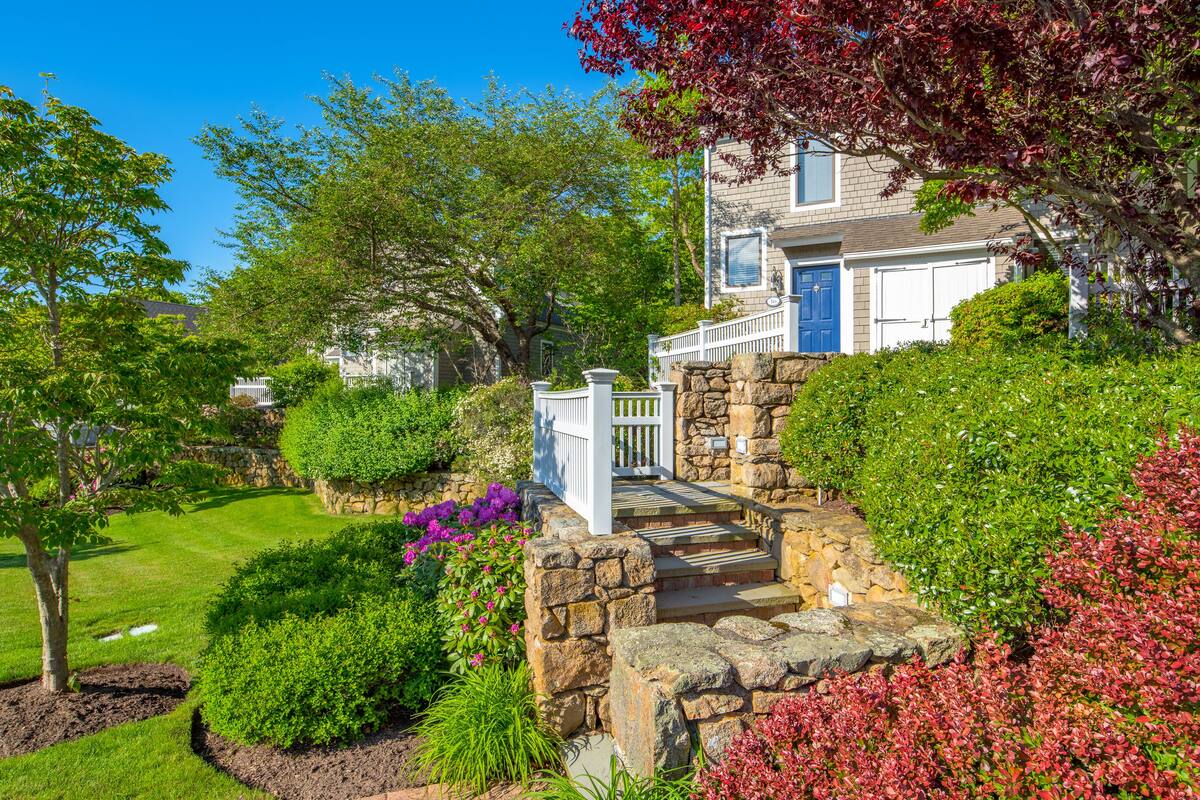 A two-story house with a stone staircase leading to a white door, surrounded by lush greenery and flowering plants.