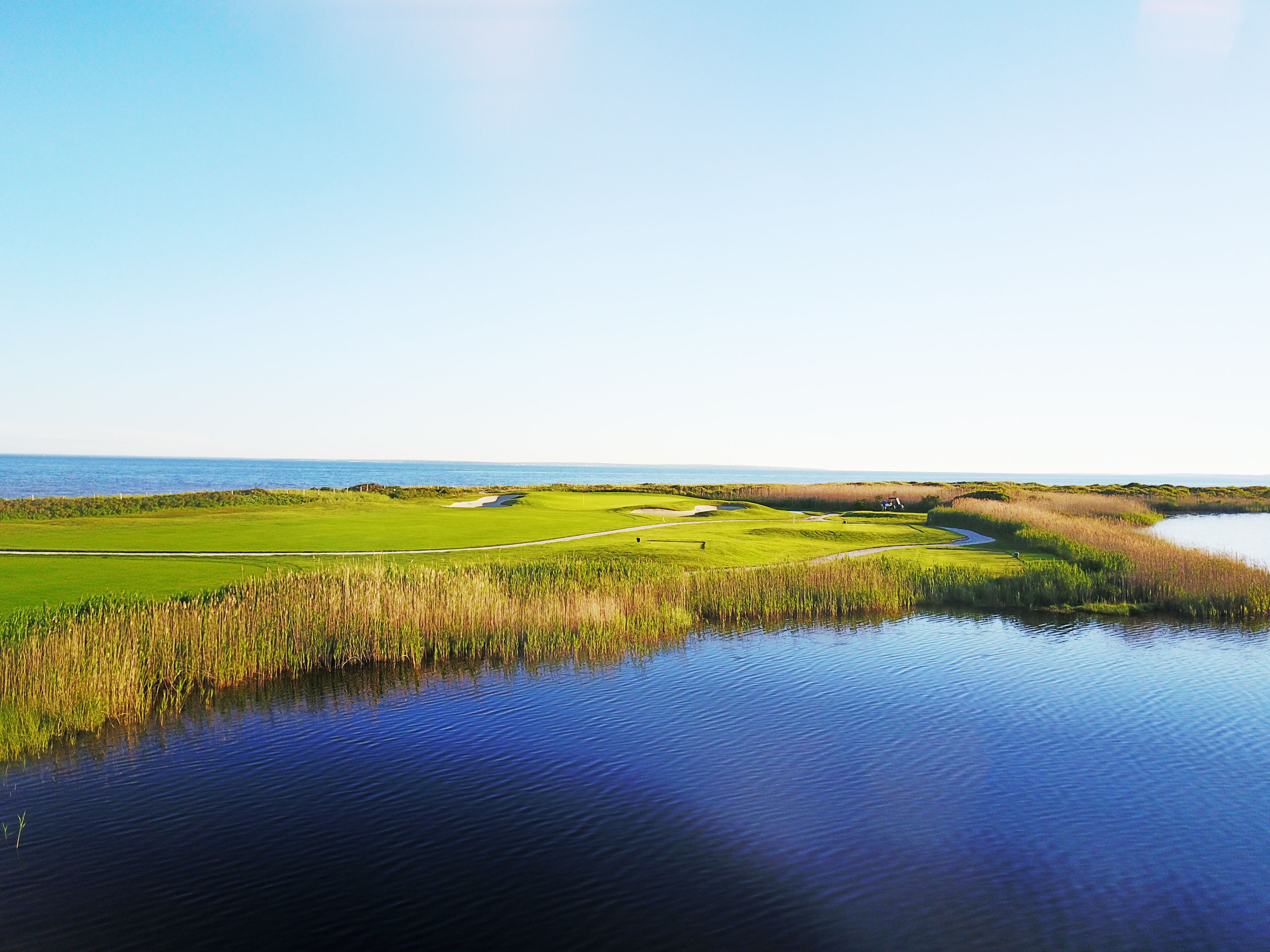 Un campo de golf con una masa de agua en primer plano y una vista despejada del océano al fondo.