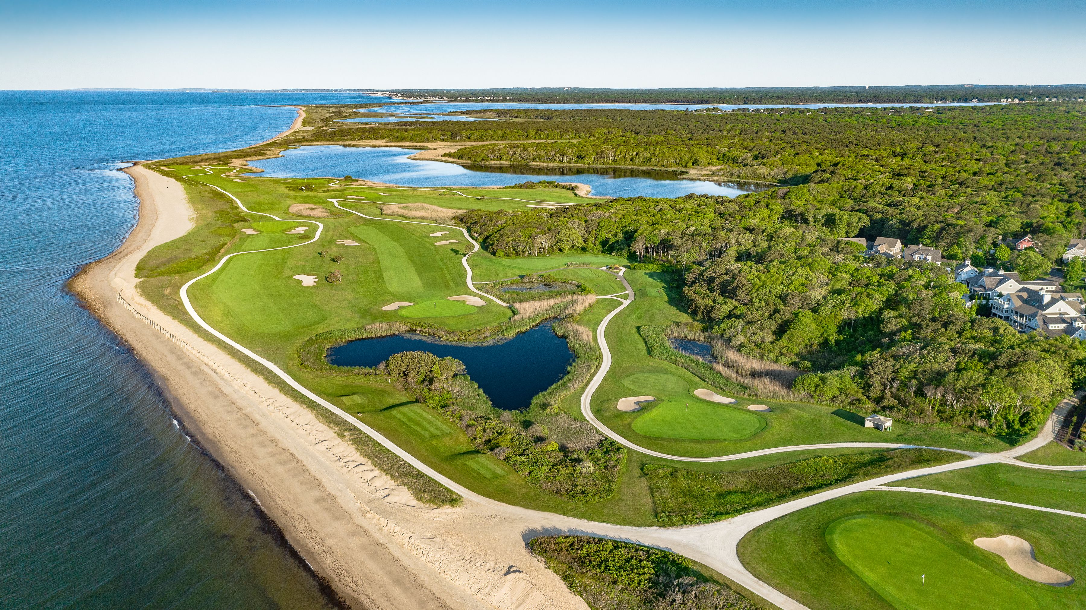 Luftaufnahme eines Golfplatzes mit Strand, Wasserflächen und bewaldeten Gebieten.