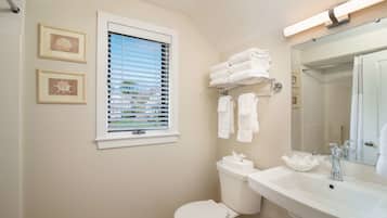 A bathroom with a toilet, sink, and window with blinds. There are towels on a rack and a framed picture on the wall.