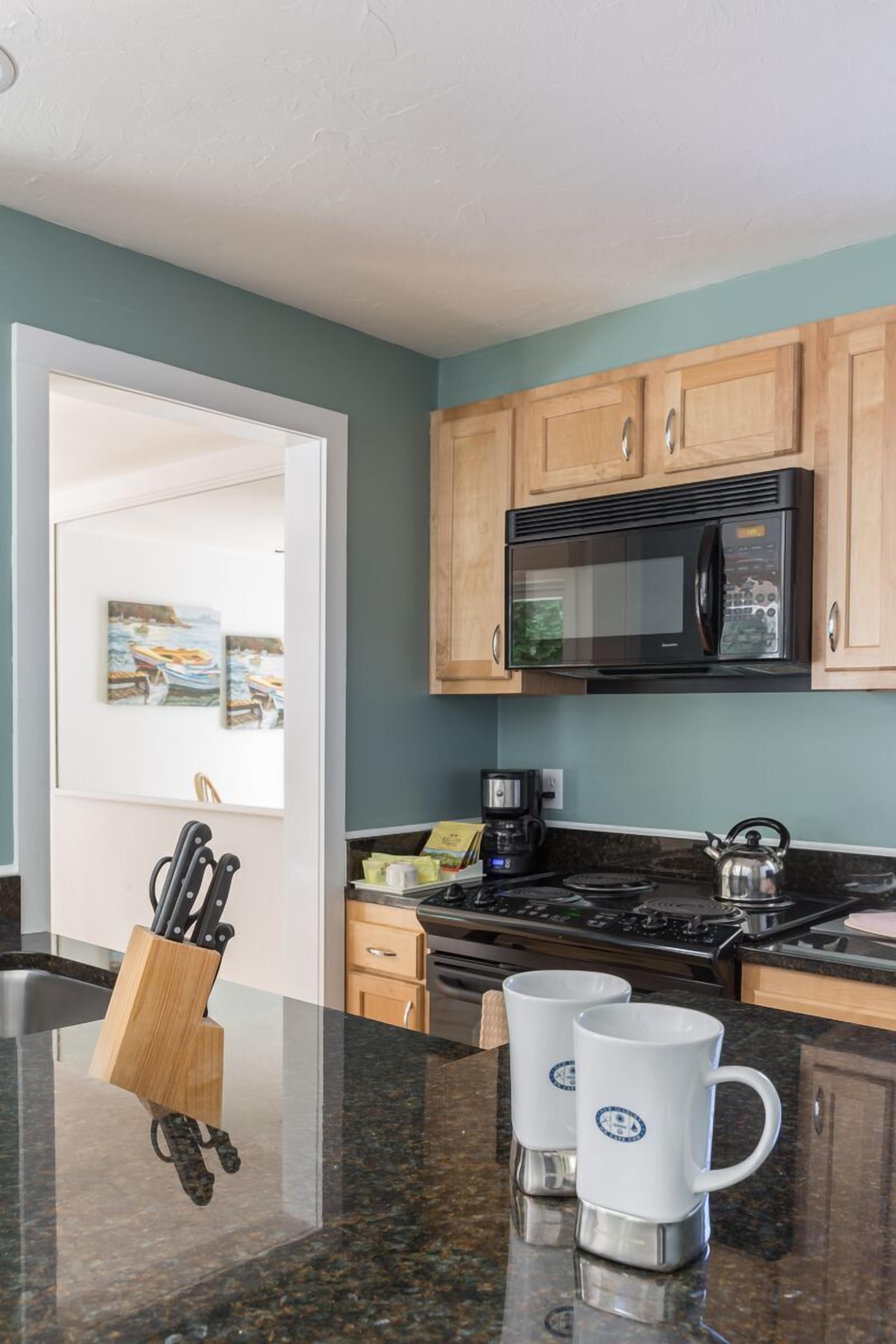 A kitchen with wooden cabinets, a black microwave, a kettle, and two white mugs on the counter.