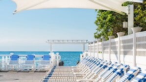Une rangée de chaises longues blanches, dotées de coussins bleus, est disposée sous un auvent blanc, offrant une vue sur la mer.