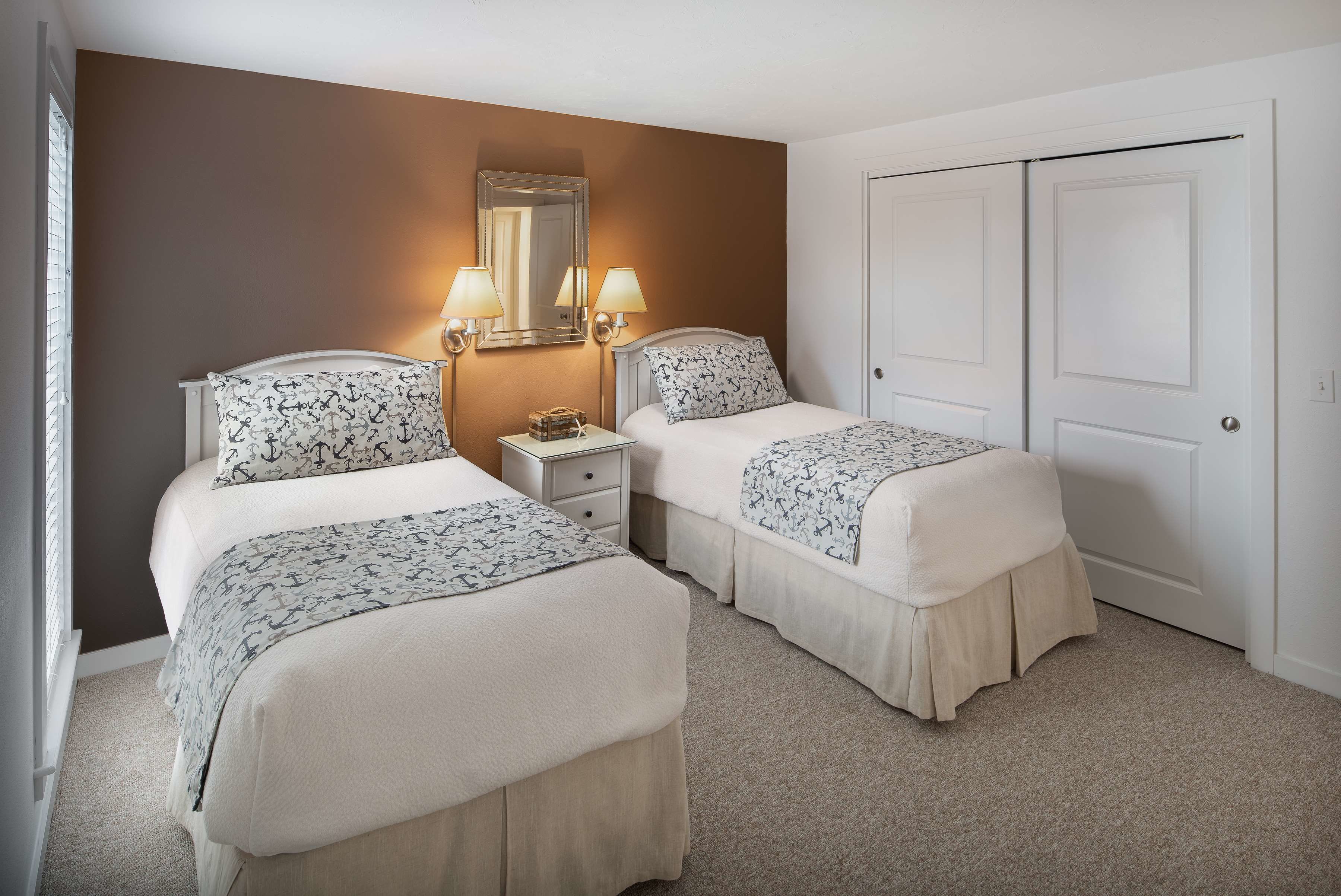 Quarto de hotel com duas camas, uma mesa de cabeceira com luminárias, um espelho e um guarda-roupa.