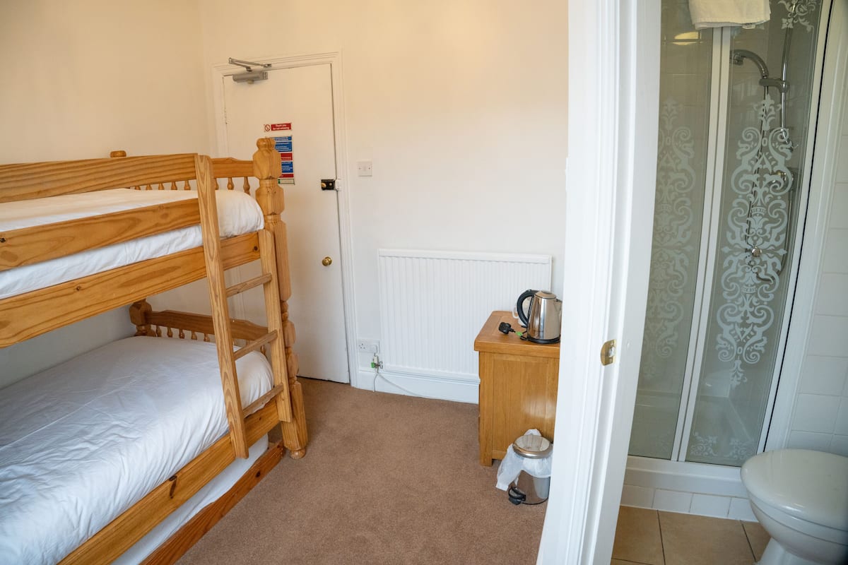 A room with three wooden bunk beds, a small wooden table with a kettle, a white radiator, and a glass shower enclosure.