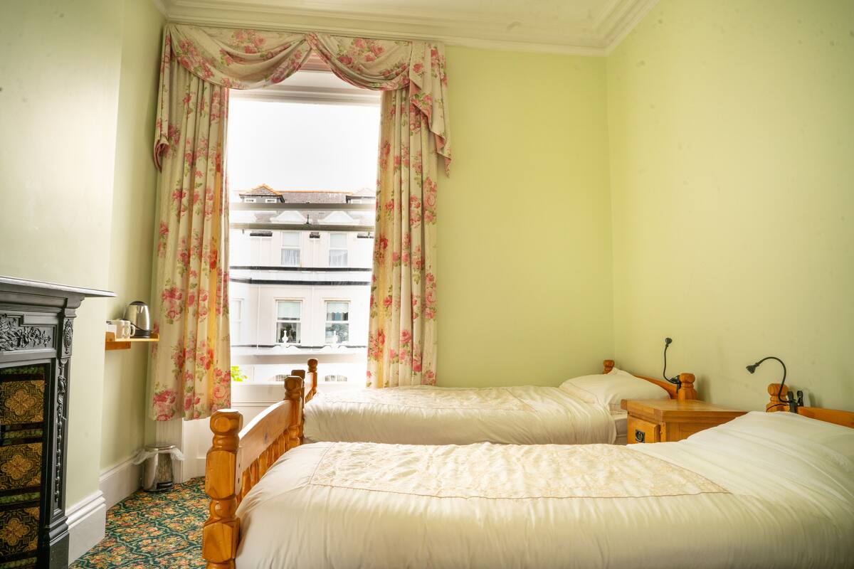 A room with two beds, a wooden headboard, a small table, and a window with floral curtains.
