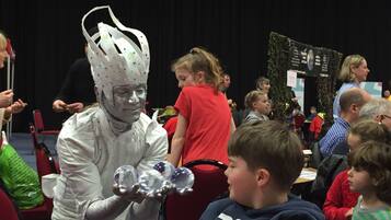 A person in a white costume holding crystal balls, interacting with children in a room with red chairs and a stage in the background.
