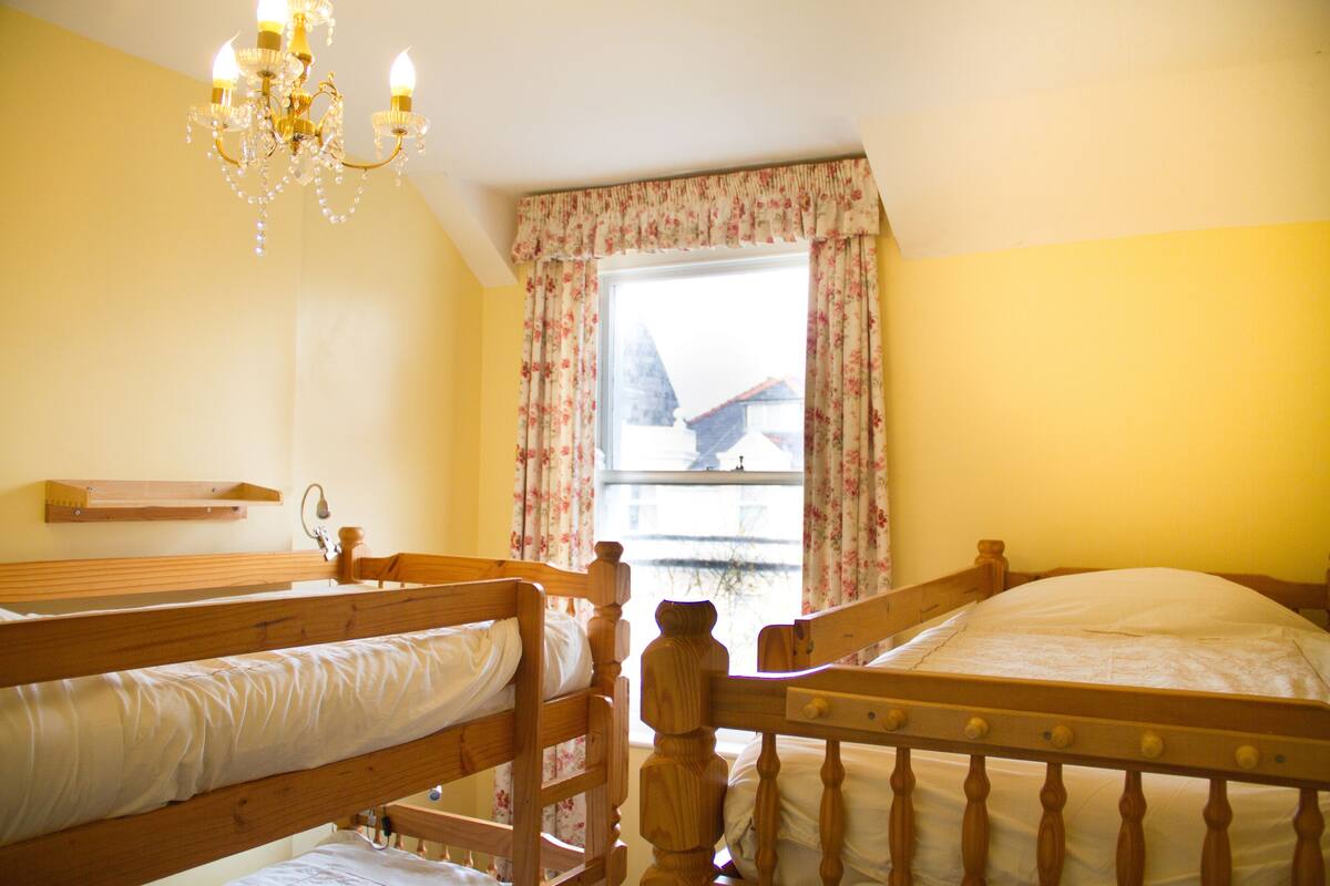 A room with two wooden beds, a chandelier, a window with floral curtains, and a wooden shelf.