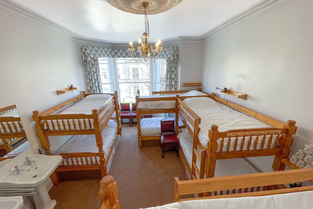 A room with four wooden bunk beds, a chandelier, a window with curtains, and a small table with a mirror.