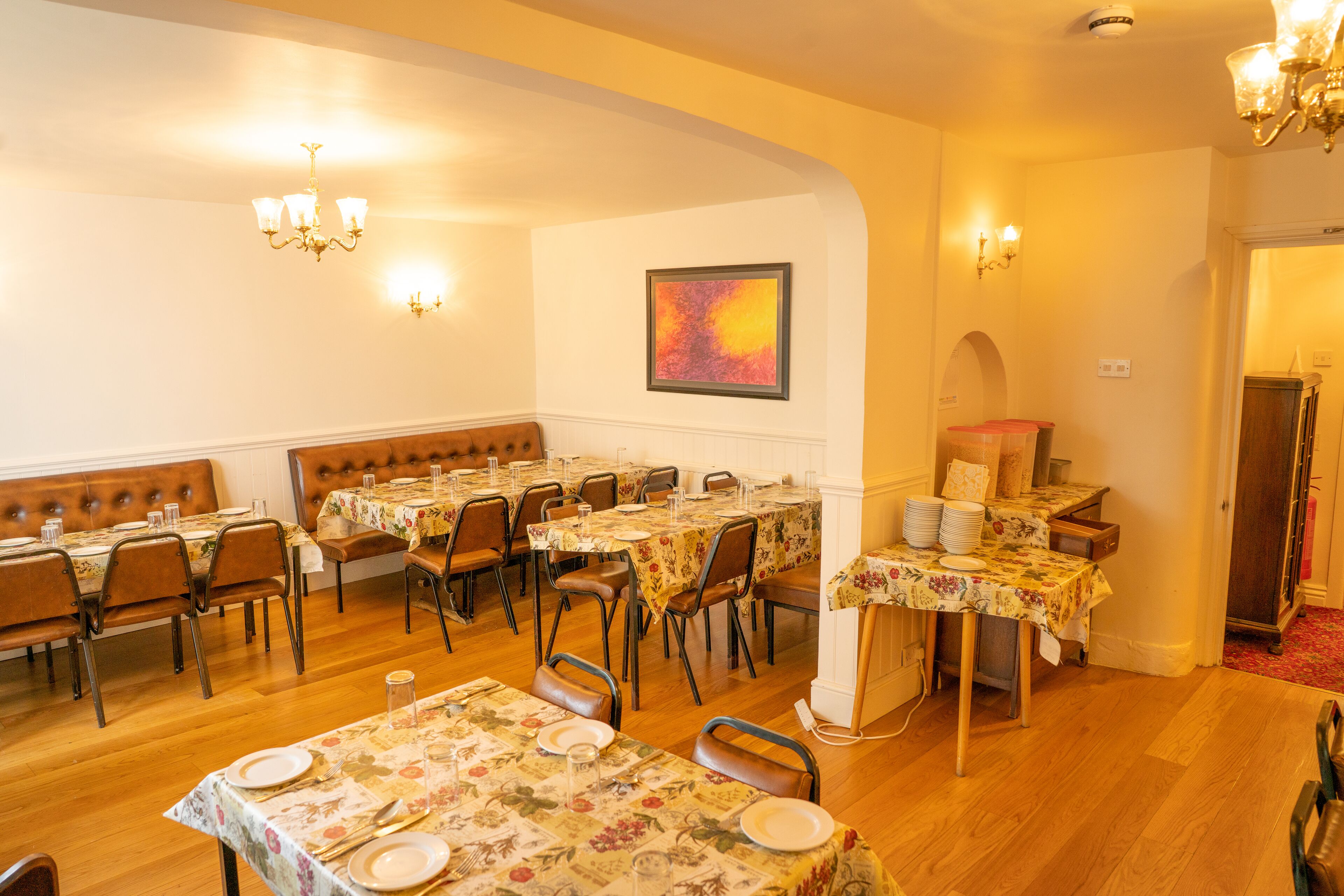 A dining area with multiple tables set for service, wooden flooring, and a chandelier.