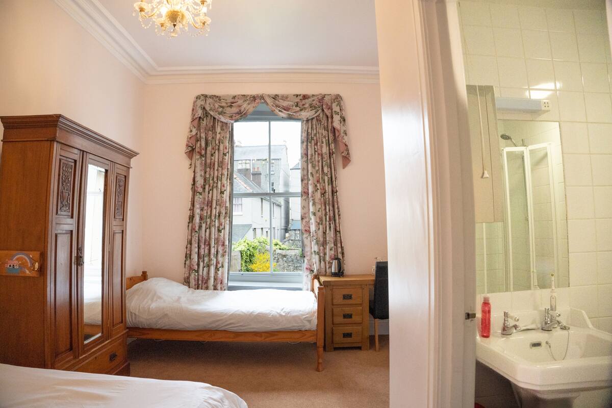 A bedroom with a bed, a wooden wardrobe, a desk with a chair, a window with floral curtains, and a bathroom with a sink and mirror.