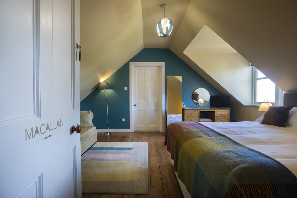 A bedroom with a sloped ceiling, a bed with a colorful blanket, a bedside table with a lamp, a dresser, and a mirror.