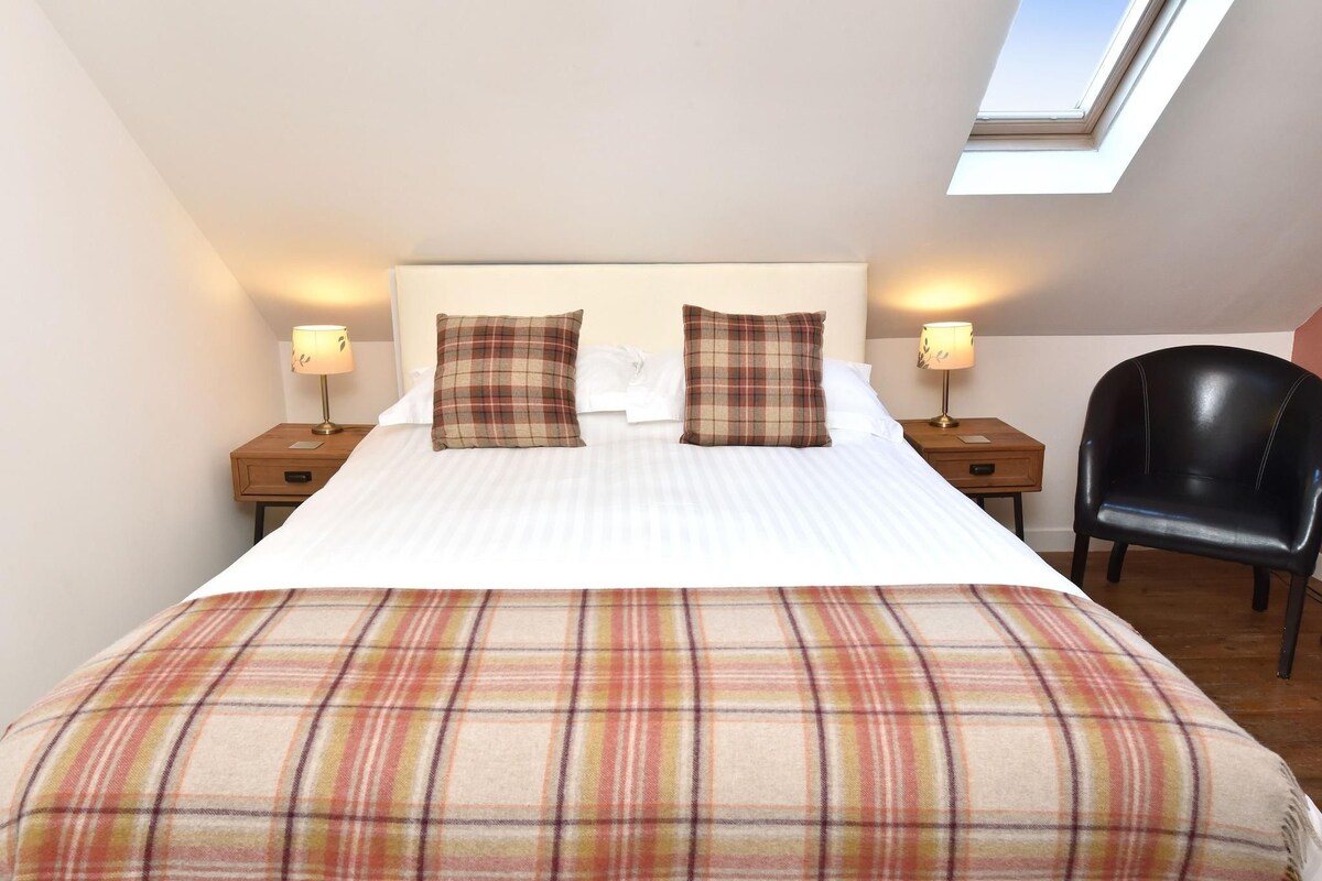 A neatly made bed with plaid bedding, two bedside lamps, a black chair, and a skylight above.
