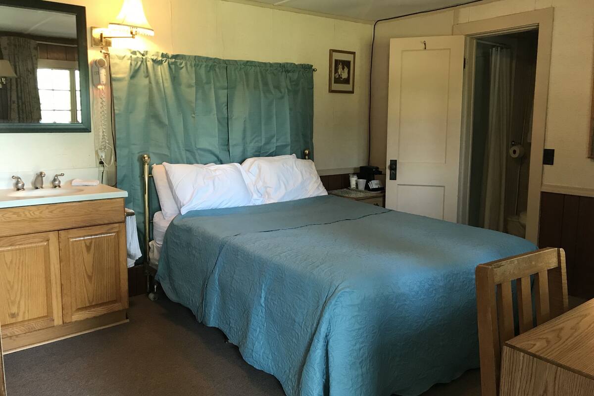 A small, cozy bedroom with a single bed, a wooden dresser, a bedside table with a lamp, and a door leading to another room.