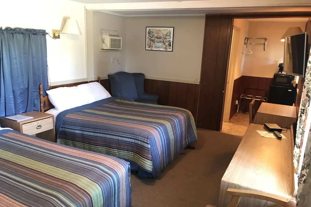 A hotel room with two beds, a desk, a chair, a small table, a lamp, a picture on the wall, and a door leading to another room.