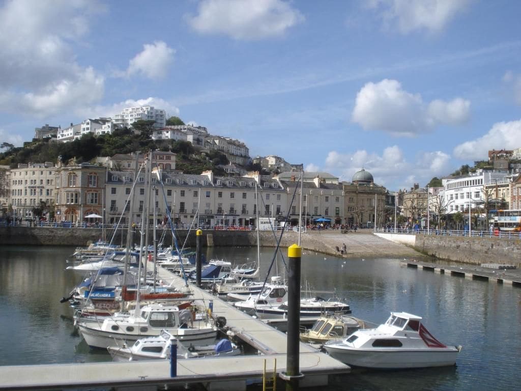 A marina with boats docked, a waterfront promenade, and buildings in the background.