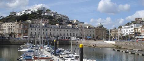 A marina with boats docked, a waterfront promenade, and buildings in the background.