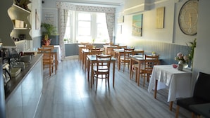 A dining area with multiple wooden tables and chairs, a kitchen area with a counter and shelves, and a large window with curtains. - Tusker Lodge (Torquay)