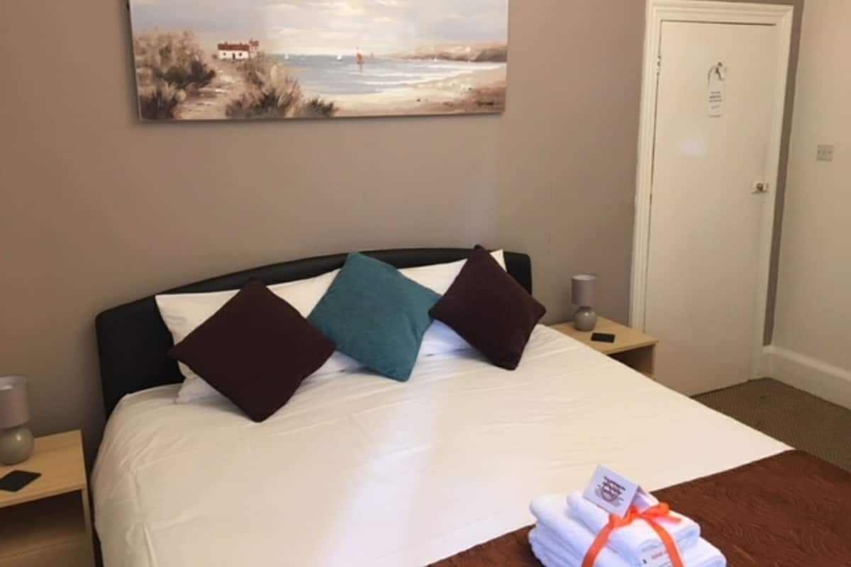 A neatly made bed with white linens and three pillows in dark and teal colors, a painting of a beach scene on the wall, and a door with a lock and a keyhole.